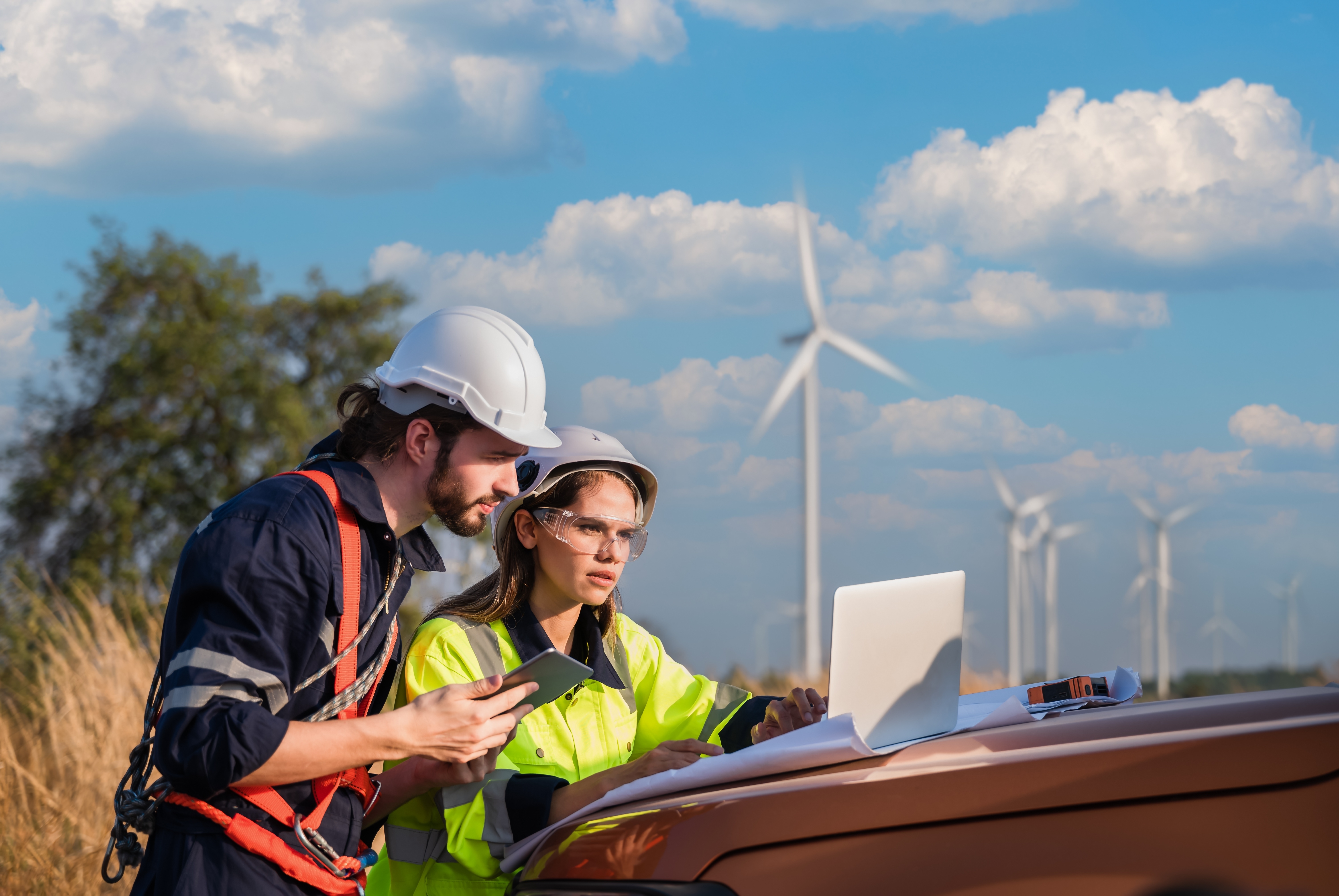 A male and a female colleague in safety gear working together on a backdrop of wind mills